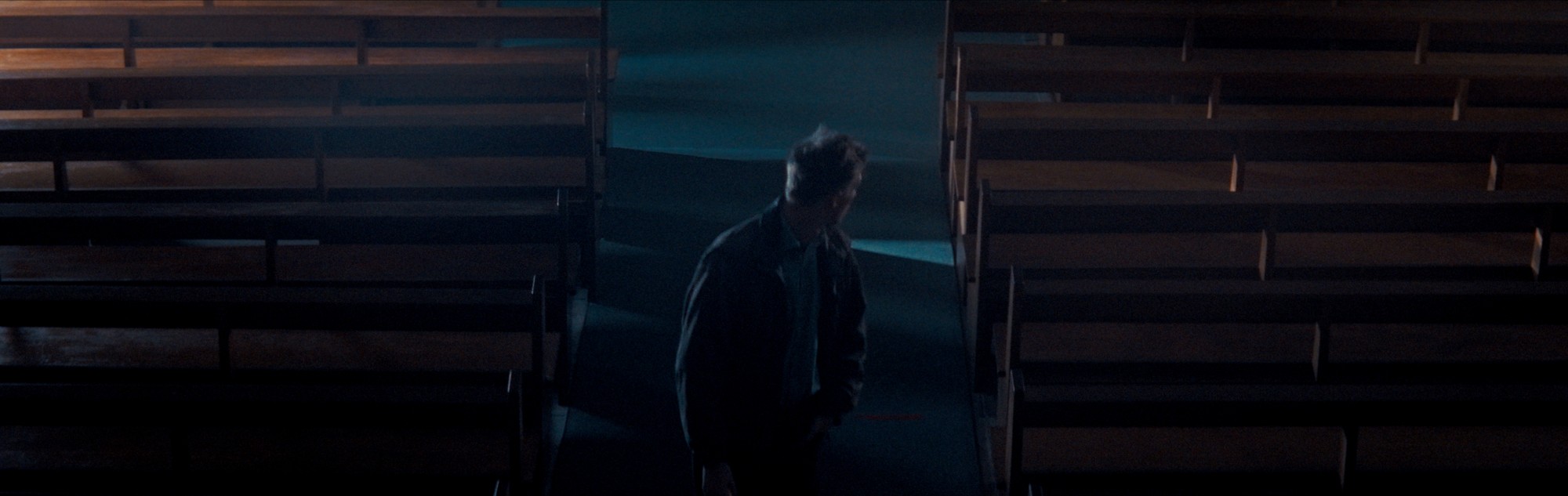 A solitary figure between rows of church pews, edged in cool light.