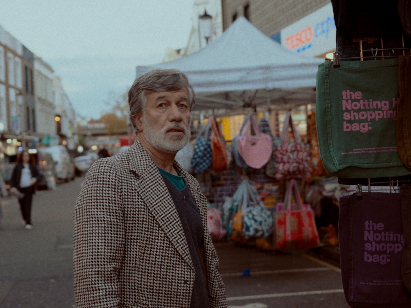 A market trader looks towards the camera beside a display of shopping bags.